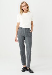 Grau gestreifte Hosen mit maßgeschneidertem Schnitt, mit seitlichen Taschen und flachem Vorderteil, kombiniert mit einem kurzärmeligen cremefarbenen Oberteil. Schwarze Schuhe.