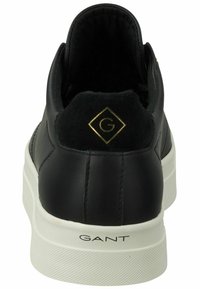 Black leather sneaker with a rounded shape, white rubber sole, and a back collar featuring a green and gold logo detail.