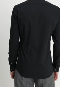 Zwarte langetermijnshirt gemaakt van gladde stof, met een gebogen zoom, knoopmanchetten en een standaard kraag. Achteraanzicht weergegeven.