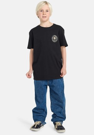 T-shirt nera a maniche corte con scollatura rotonda e patch del logo sul petto, abbinata a jeans in denim blu a gamba larga e sneaker nere.