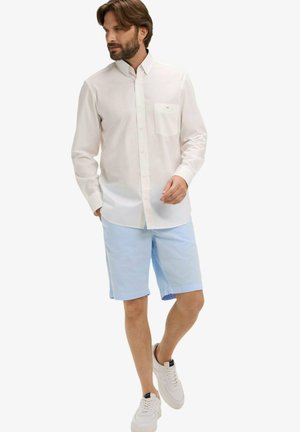 Homme portant une chemise blanche à manches longues boutonnée, un short bleu clair et des baskets blanches, marchant avec une main dans la poche.
