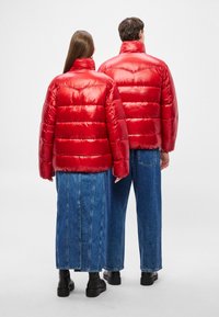 Κόκκινα γυαλιστερά μπουφάν τύπου puffer με οριζόντια καπιτονέ ραφή και ψηλούς γιακάδες, φορεμένα πάνω από μακριές μπλε φούστες από τζιν, σε συνδυασμό με μαύρα μποτάκια αστραγάλου.
