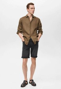 Braune Leinenhemd mit Knöpfen, dunkle Denim-Shorts mit umgeschlagenen Säumen und schwarze Sandalen mit Riemen. Glatte Textur, lässige Passform, neutrale Farbpalette.