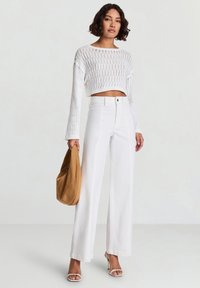 Top corto bianco all'uncinetto con maniche lunghe, abbinato a pantaloni bianchi a gamba larga a vita alta. Borsa in suede marrone e sandali con tacco chiari.