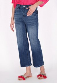 Pantalones de mezclilla de pierna ancha en azul oscuro con cintura alta y detalles desgastados. Combinados con sandalias planas que tienen borlas en crema y rojo, junto con acentos decorativos.