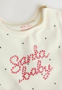 Cremefarvet babydragt med røde stribede broderede tekst, der står "Santa baby", og små sorte hjerte mønstre. Inkluderer trykknaplukninger ved skuldrene.