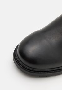 Μαύρη δερμάτινη μπότες με λείο, στρογγυλό άκρο, διαθέτοντας υφή στη σόλα και ανάγλυφες ραφές κατά μήκος της άκρης για πρόσθετη αντοχή.