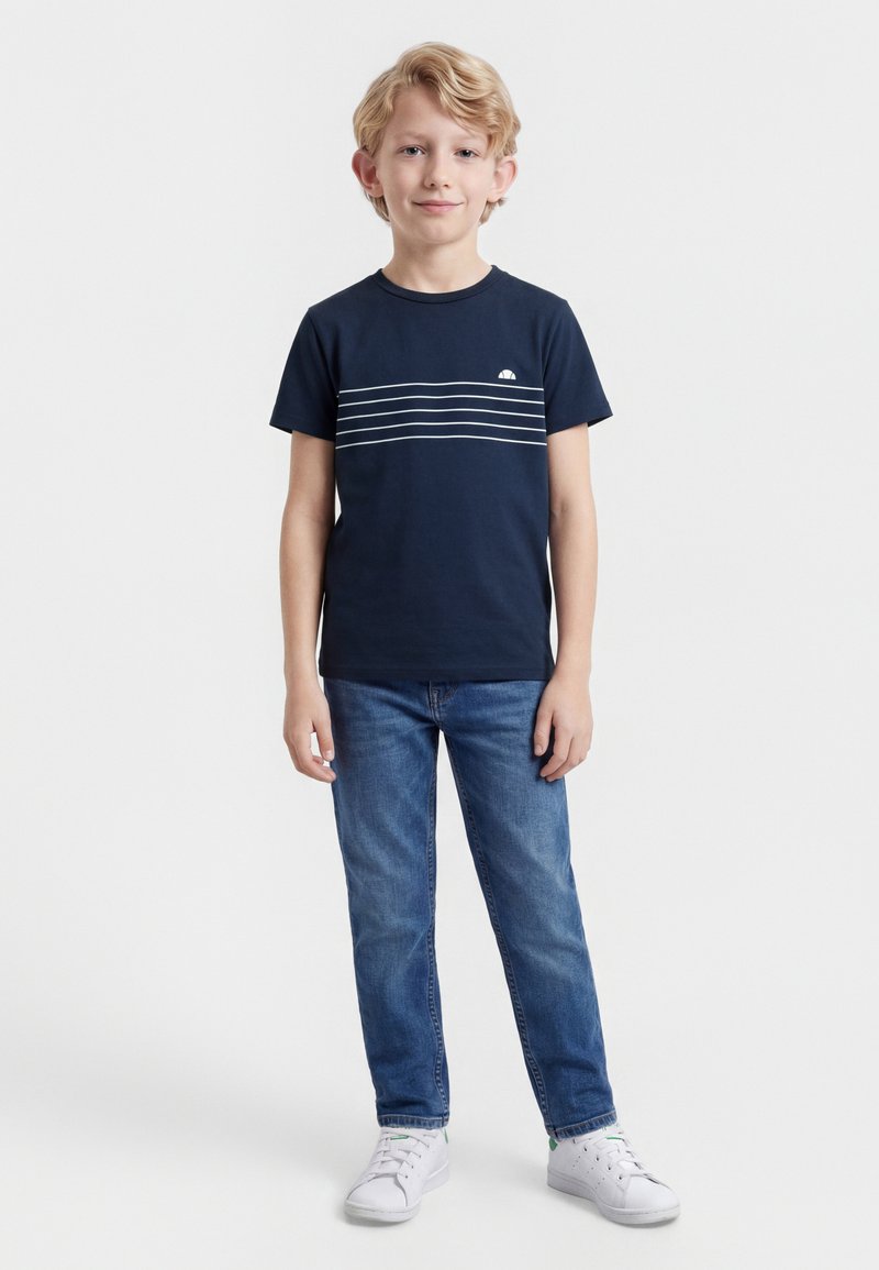 Αγόρι με ξανθά μαλλιά στέκεται στραμμένο προς τα εμπρός, φοράει μπλε ναυτικό ριγέ T-shirt, μπλε τζιν και λευκά αθλητικά παπούτσια σε άσπρο φόντο.