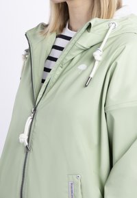 Ljusgrön regnjacka med vita dragskor, silverfärgad dragkedja och paraplylogga, bärs över en svart och vit randig skjorta.