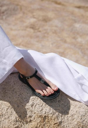 Czarny sandał z paskiem w kształcie litery T, ozdobiony okrągłym srebrnym elementem, z płaską podeszwą i teksturowanymi paskami z tkaniny, umieszczony na jasnej kamiennej powierzchni.