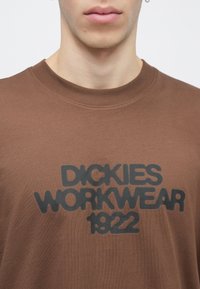 Κοντινή εικόνα ενός ατόμου που φοράει καφέ μπλούζα με στρογγυλή λαιμόκοψη και μαύρο κείμενο "Dickies Workwear 1922" στο στήθος.