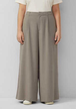 Personne portant un pantalon large beige et des baskets blanches, debout devant un fond gris clair.