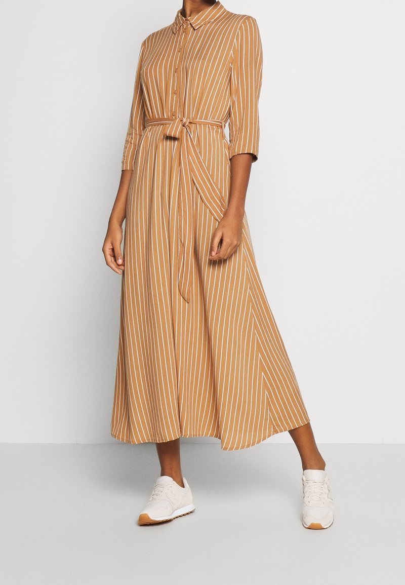 Femme portant une robe midi boutonnée à rayures verticales beige et blanches, avec une ceinture nouée et des baskets blanches, se tenant contre un mur uni.