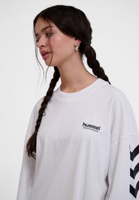 Sötét fonott hajjal rendelkező nő ezüst karika fülbevalóval és fehér Hummel Sportswear hosszú ujjú pólóval, melyen fekete csíkos minta van az ujjon.