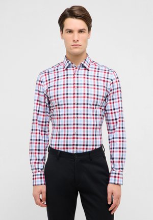 Mann trägt ein enganliegendes, langärmliges Hemd mit Knöpfen und rotem, blauem und weißem Kariertmuster sowie schwarze Hose, steht vor einem einfarbigen Hintergrund und schaut nach vorne.