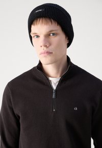 Čierna rebrovaná čiapka a tmavý textúrovaný zipsový sveter s malým logom. Model má svetlú pleť a krátke vlasy.