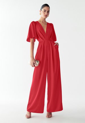 Žena v červeném overal s širokými nohavicemi a klouzavými rukávy, která drží zlatou kabelku, stojí proti jednoduchému bílému pozadí.