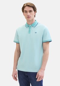 Polo shirt van lichtblauwe stof met een kraag, een knoopsluiting met drie knopen en contrasterende manchetten. Voorzien van een klein logo op de borst.