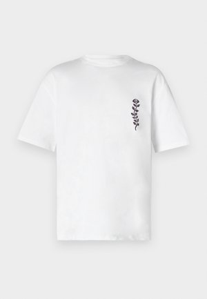 T-shirt en coton blanc à manches courtes et col rond. Présente un petit motif floral noir sur le côté gauche de la poitrine.
