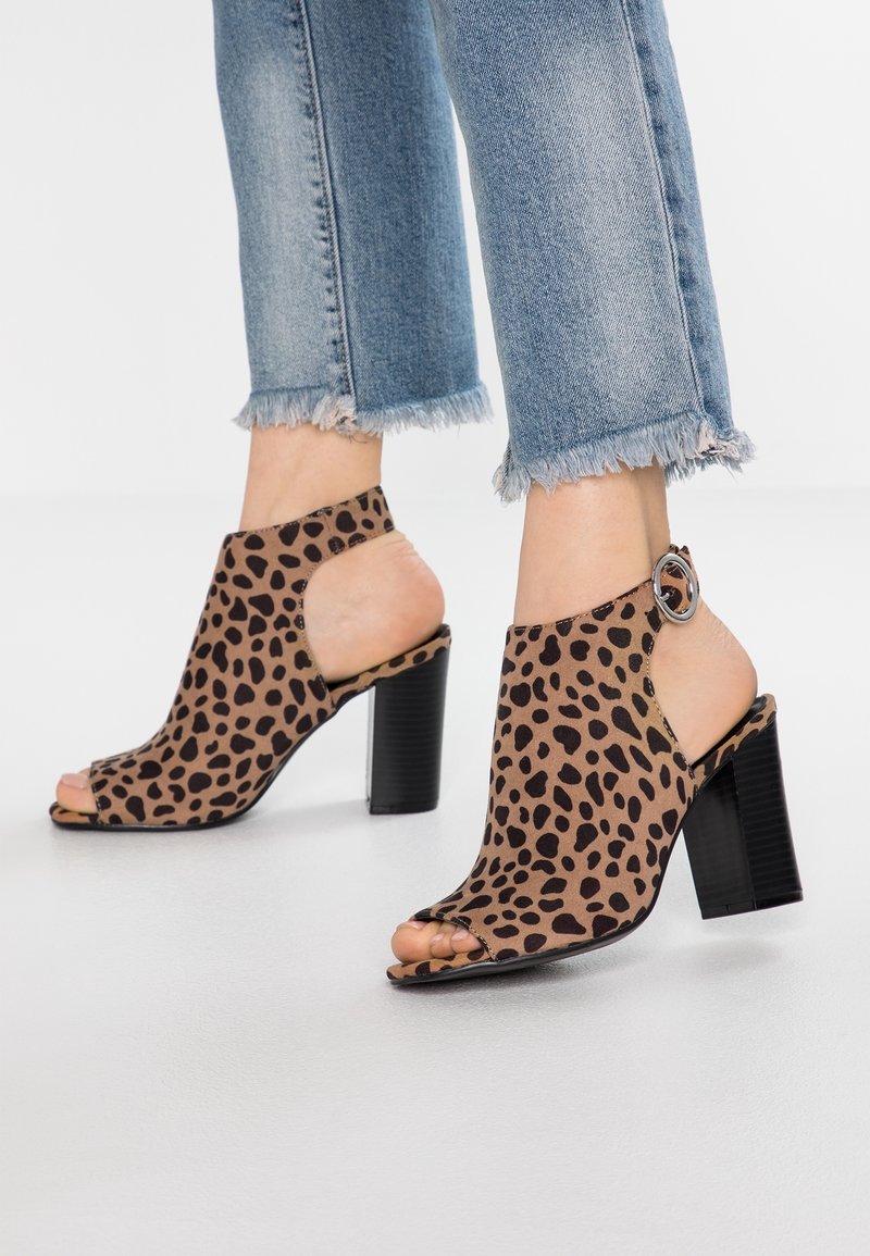 Sandales à talons hauts ouvertes à l'avant, marron et noires tachetées, avec une bride à la cheville, portées avec un jean bleu clair à ourlet effiloché.