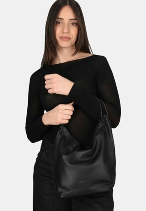 Giovane donna con capelli castani lisci che indossa un top e pantaloni neri, mentre tiene una grande borsa in pelle nera su sfondo bianco.