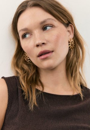 Mladá žena so svetlohnedými vlasmi má na sebe tmavý textúrovaný tielko bez rukávov a zlaté náušnice v tvare slnka, pozrie sa mierne do strany.