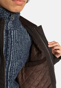 Veste extérieure marron avec une fermeture éclair et doublure matelassée marron. Pull bleu marine en dessous avec des rayures verticales bleu clair et blanches.