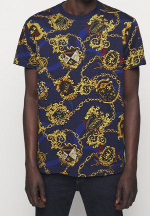 Mann trägt ein marineblaues T-Shirt mit aufwendigen goldenen Ketten- und Wappenmustern, kombiniert mit dunklen Jeans, vor einfachem hellem Hintergrund.