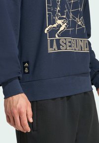 Sudadera azul marino con diseño gráfico dorado que incluye un jugador, texto "LA SEGUNDA" y etiqueta de marca Adidas en el dobladillo. Tejido suave y texturizado.