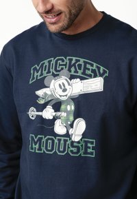 Sudadera de color azul marino con un Mickey Mouse vintage que lleva una bufanda de cuadros, cargando una tabla de madera y un bastón de esquí, con el texto "MICKEY MOUSE" en verde y negro.