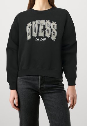 Osoba ubrana w czarną bluzę z napisem „GUESS Est. 1981” dużymi białymi literami z konturem, w połączeniu z ciemnymi dżinsami.