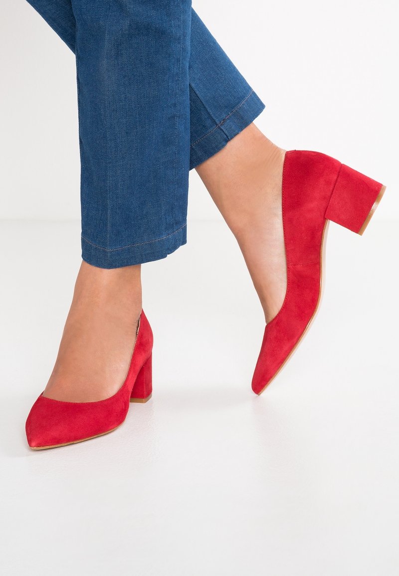 Femme portant un jean court bleu et des chaussures rouges en daim à talons blocs, un pied à plat sur le sol et l'autre sur la pointe des pieds.