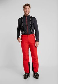 Leende man iförd svart zip-up jacka, röda skidbyxor med svarta hängslen och svarta skidstövlar står framför en enkel grå bakgrund.