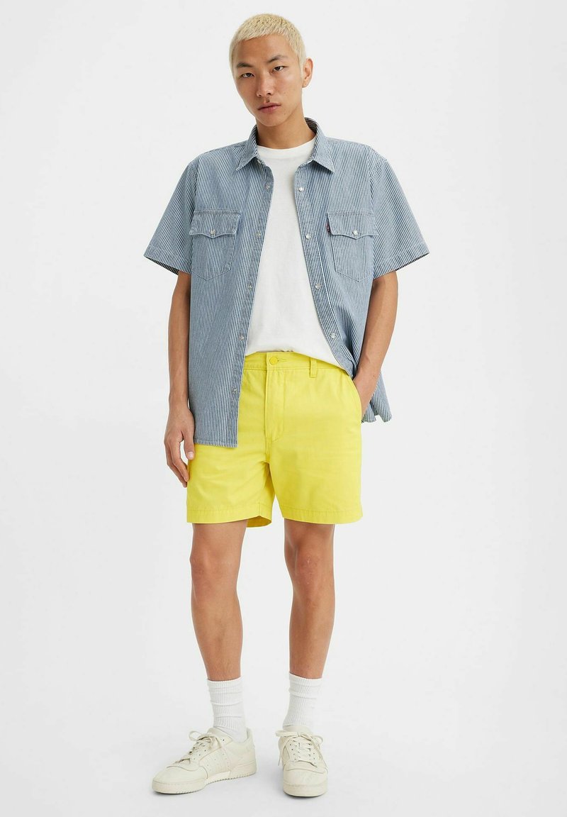 Randig kortärmad skjorta i blått och vitt, tillsammans med ljusgula shorts. Vit t-shirt under. Neutrala sneakers och strumpor.