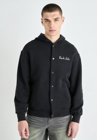 Svart hettegenser med trykknapper, med "Dark Side" tekst brodert på brystet. Myk stoff, avslappet passform, minimal tekstur.