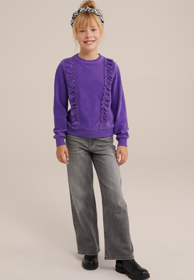 Vijolična pletena jopica z nagubanim detajlom obeh straneh, v kombinaciji z sivo širokima hlačami in črnimi gležnjarji. Črno-bela naglavna trak.