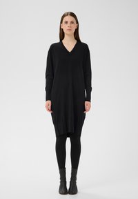 Schwarzes, langärmeliges Strickkleid mit V-Ausschnitt, das eine lockere Passform, einen gerippten Saum und eine asymmetrische Länge aufweist, kombiniert mit schwarzen Leggings und Stiefeletten.