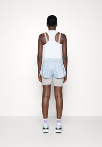 Žena stojící zády, oblečená v bílém tílku, světle modré vzorované sukni přes šedé šortky, pruhovaných ponožkách a světle modrých teniskách na bílém pozadí.