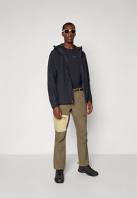 Mann iført svart jakke, svart skjorte, beige bukser med en lys beige lapp, svarte sko og solbriller med hvit innfatning mot ensfarget bakgrunn.