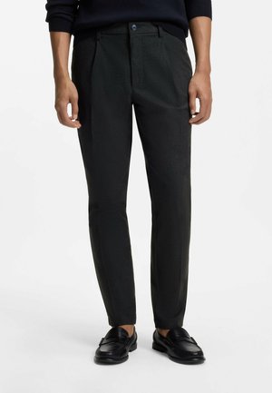 Mann, der schwarze maßgeschneiderte Hosen und schwarze Loafers trägt, steht vor einem schlichten hellen Hintergrund mit entspannt an den Seiten hängenden Händen.