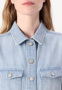 Lyseblå denimjakke med spiss krage, sølvfargede knapper, og to brystlommer med knappeklaffer. Subtil tekstur og sømdetaljer.