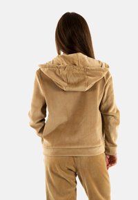 Felpa in corduroy beige con cappuccio, texture a coste e polsini aderenti. Abbinata a pantaloni in corduroy dello stesso colore, caratterizzati da un aspetto morbido e confortevole.