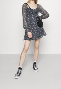Modré květinové mini šaty s nabíranými rukávy, volánkovým lemem a přiléhavým korzetem; doplněné černými vysokými teniskami a malou černou křížovou taškou.