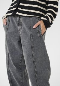 Laisvai parinkti pilki džinsai su minkšta tekstūra, turi šonines kišenes ir matomas siuvimo detales, derinami su dryžuotu megztiniu.