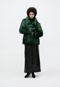 Πράσινο παλτό από συνθετική γούνα με ψηλό γιακά, μακριά μανίκια και κούμπωμα στο μπροστινό μέρος, συνδυασμένο με στενή μαύρη δερμάτινη φούστα και μαύρες γόβες.