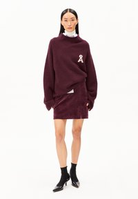Bordeauxfarbener strukturierter Pullover mit einem A-förmigen Patch, kombiniert mit einem passenden figurbetonten Rock. Das Model trägt schwarze Absatzstiefeletten für den Kontrast.