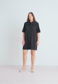 Black denim shirt dress with short sleeves, front pockets, and button closure. Model wearing white heeled sandals stands against a light background.