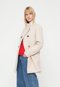 Cappotto in misto lana beige con colletto alto, chiusura anteriore a bottoni e due grandi tasche. Tessuto testurizzato completato da bottoni scuri.