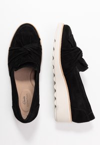 Mocassins noirs en daim à enfiler avec un détail de nœud torsadé et une épaisse semelle blanche, présentés de vue de dessus et de côté sur un fond blanc.