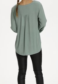 Haut vert clair à manches longues avec un col rond, détails de pli dans le dos, texture douce et ourlet arrondi, porté avec un pantalon en cuir noir.